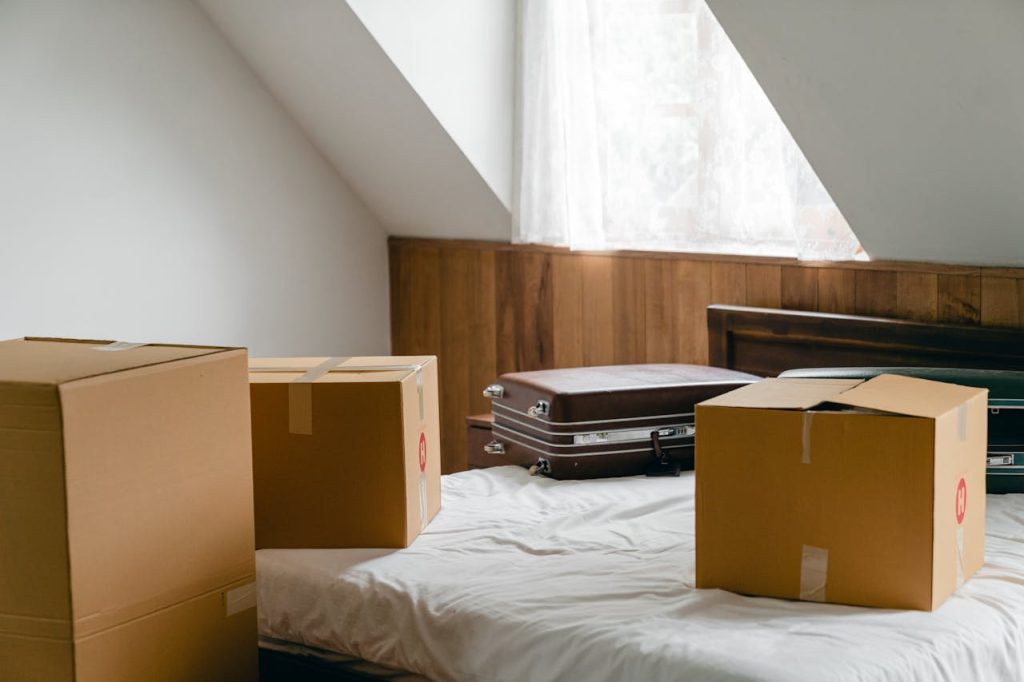 Small cardboard boxes and leather cases placed on bed with sheet near big box in light room of house under sloping roof